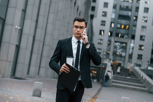 Walking and talking by phone. Businessman in black suit and tie is outdoors in the city. - Foto, Imagen