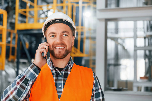 Talking by phone. Construction worker in uniform is in the factory. - Foto, afbeelding