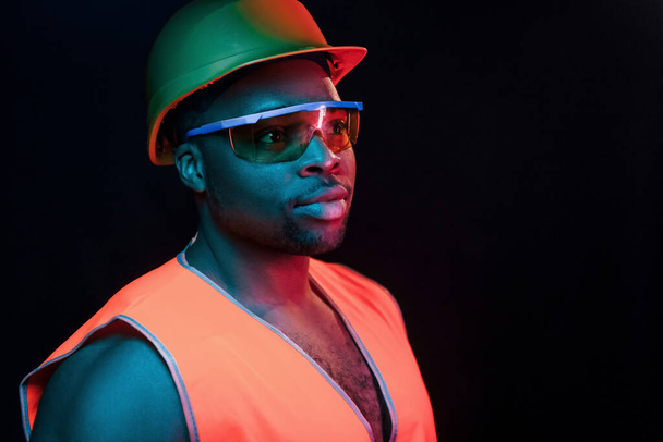 Construction worker in uniform and hard hat. Futuristic neon lighting. Young african american man in the studio. - Фото, зображення