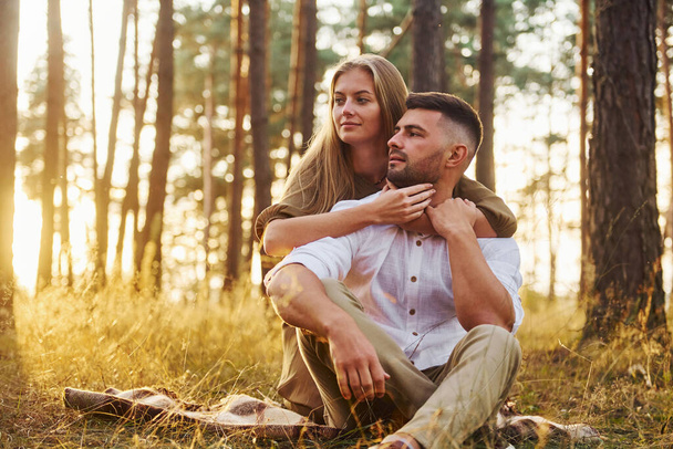 Sitting on the ground. Happy couple is outdoors in the forest at daytime. - Фото, изображение