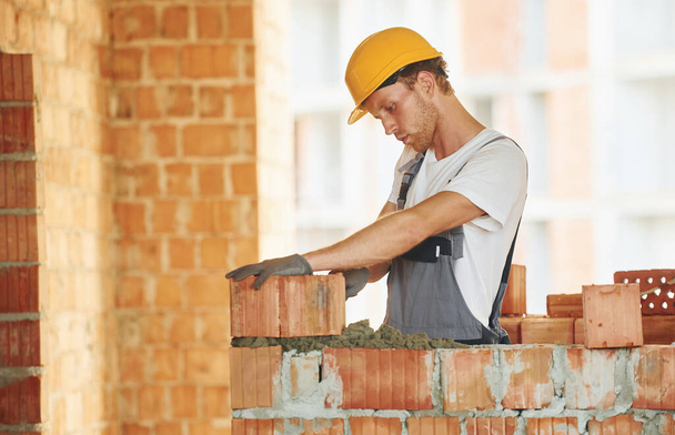 Bricks installation. Young man working in uniform at construction at daytime. - Zdjęcie, obraz