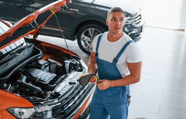 Orange colored car. Man in uniform is repairing broken automobile indoors. - Foto, Imagen