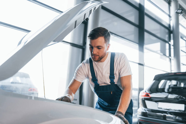 Quality service. Young man in white shirt and blue uniform repairs automobile. - Fotó, kép