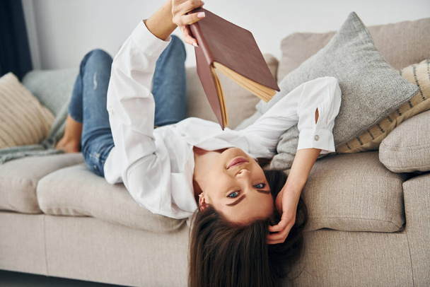 With book. Beautiful young woman in jeans and white shirt is at home. - Foto, afbeelding