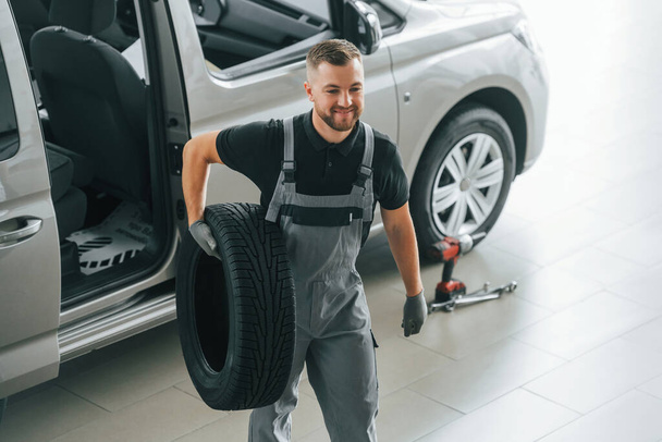 Man in uniform is working in the autosalon at daytime. Holding tire in hands. - Zdjęcie, obraz