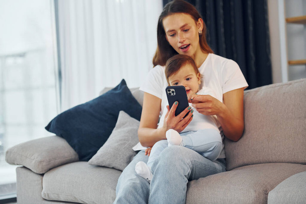With smartphone in hand. Mother with her little daughter is at home together. - Valokuva, kuva