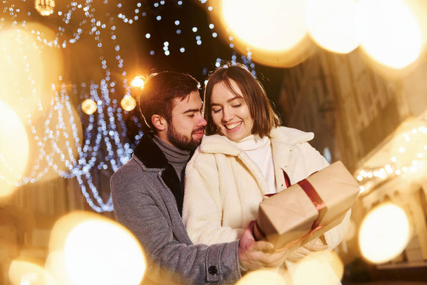 Woman holds gift box. Happy young couple celebrating New year outdoors on the street. - Photo, image