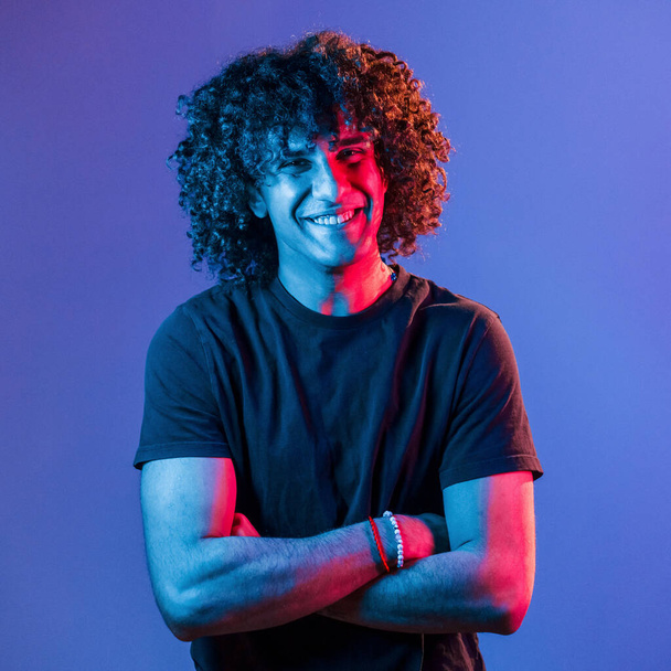 Posing for a camera. Young beautiful man with curly hair is indoors in the studio with neon lighting. - Foto, Imagem