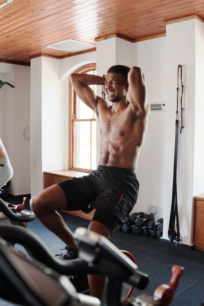 Shirtless young black man with hands behind head doing intense cardio workout on exercise bike at the gym - Fotografie, Obrázek