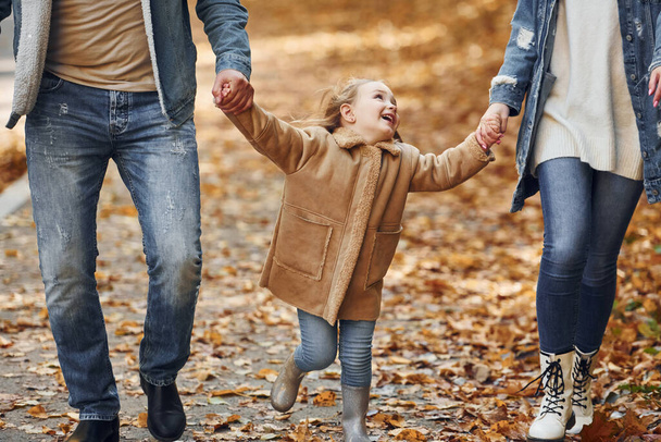 Holding daughter by hands. Happy family is in the park at autumn time together. - Foto, afbeelding