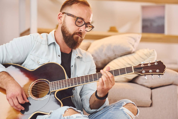 Sitting near the sofa. Man in casual clothes and with acoustic guitar is indoors. - Foto, afbeelding