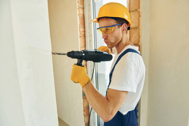 Holding drill. Young man working in uniform at construction at daytime. - Foto, imagen