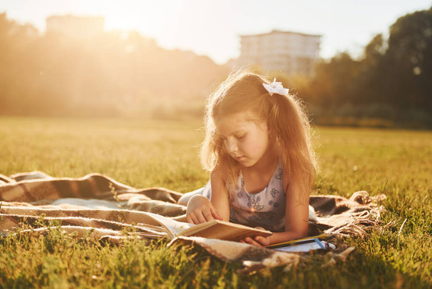 Little girl is reading book while laying down on the grass of field outdoors. - Foto, afbeelding