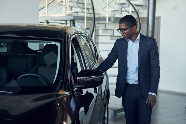 Looking at car. Young african american businessman in black suit is the autosalon. - Photo, image