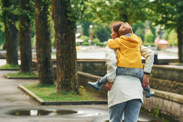 Boy is on the shoulders. Mother with her son is having a walk outdoors in the park after the rain. - Foto, afbeelding