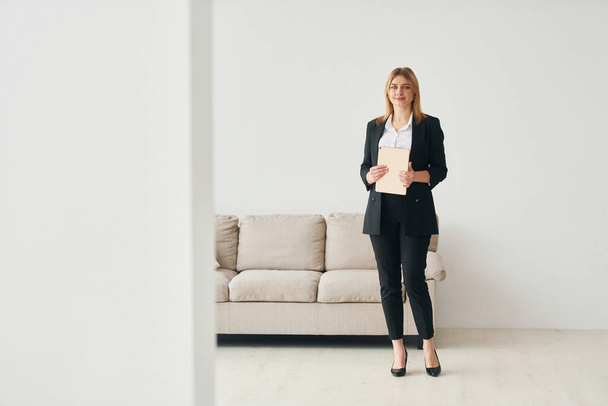 Woman in formal clothes standing near sofa indoors against white wall. - Photo, Image