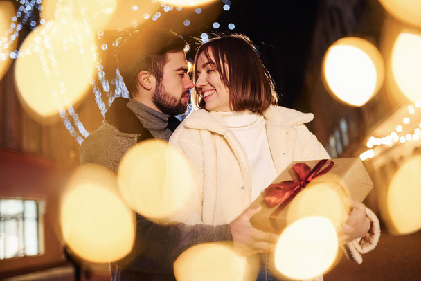 Woman holds gift box. Happy young couple celebrating New year outdoors on the street. - Foto, Imagen