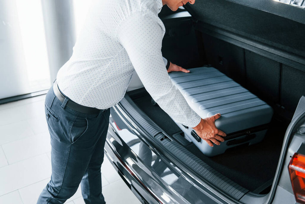 Conception of tourism. Man in formal clothes is with luggage indoors near the car. - Фото, изображение