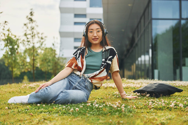 Listens to the music in headphones. Young asian woman is outdoors at daytime. - Фото, изображение