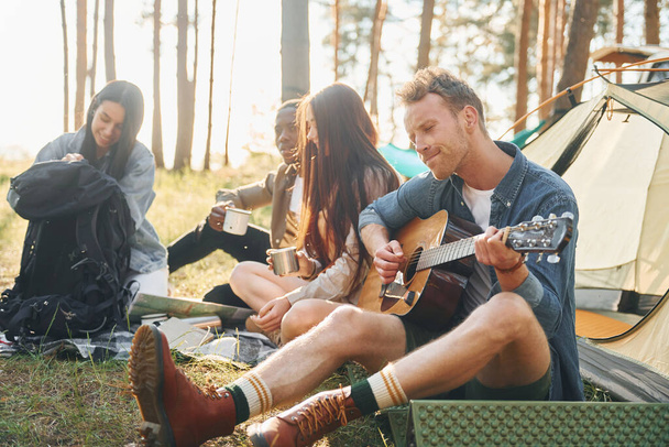 Man plays guitar. Group of young people is traveling together in the forest at daytime. - Fotografie, Obrázek
