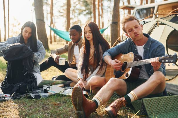 Man plays guitar. Group of young people is traveling together in the forest at daytime. - 写真・画像