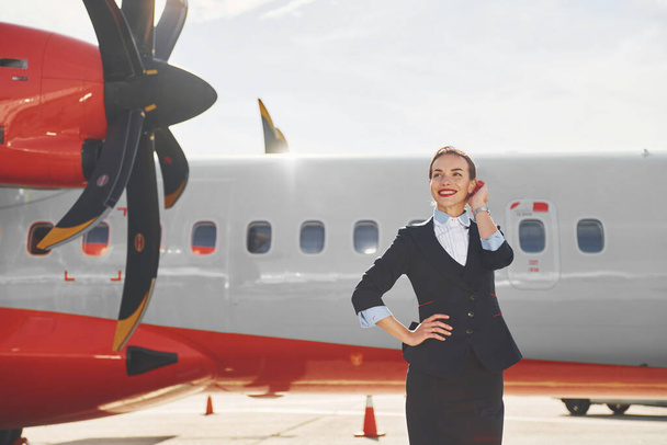 Looking far away. Young stewardess that is in formal black clothes is standing outdoors near plane. - Foto, immagini