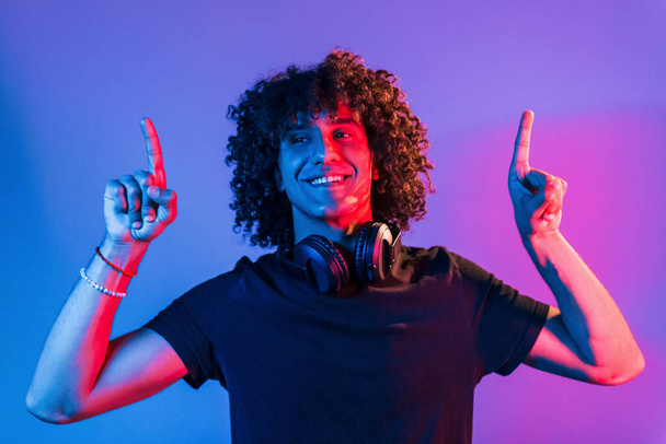 With headphones. Young beautiful man with curly hair is indoors in the studio with neon lighting. - Фото, зображення