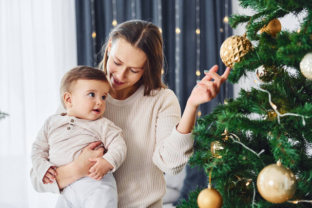 Decorating Christmas tree. Mother with her little daughter is indoors at home together. - Foto, Imagen