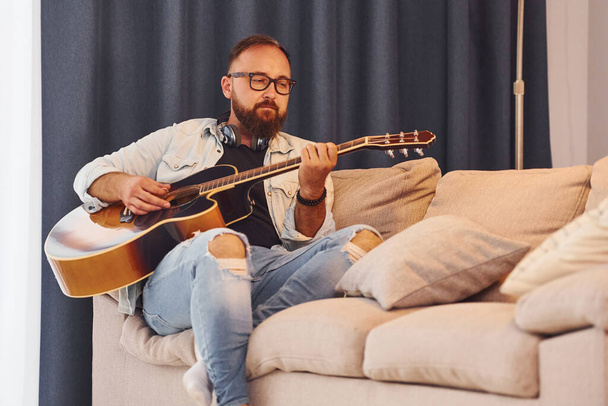 Feeling relaxed. Man in casual clothes and with acoustic guitar is indoors. - Fotoğraf, Görsel
