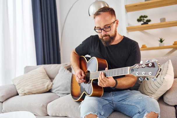 Playing the instrument. Man in casual clothes and with acoustic guitar is indoors. - Foto, Bild