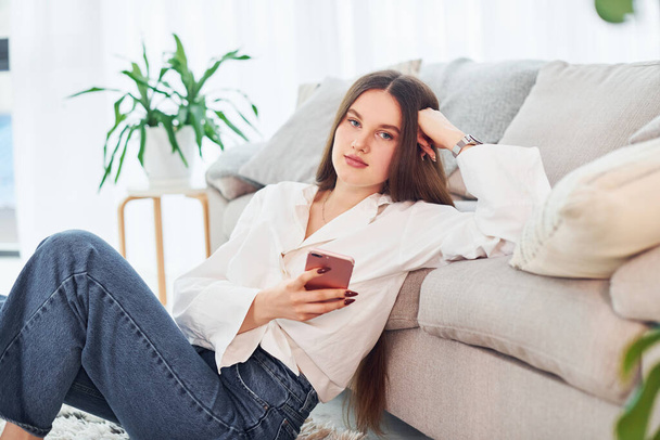 Sits near sofa with phone in hands. Young woman in white shirt and jeans is at home. - 写真・画像