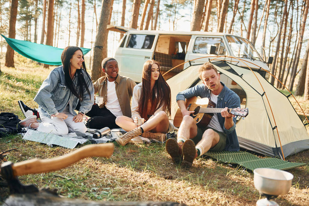 Man plays guitar. Group of young people is traveling together in the forest at daytime. - Foto, imagen