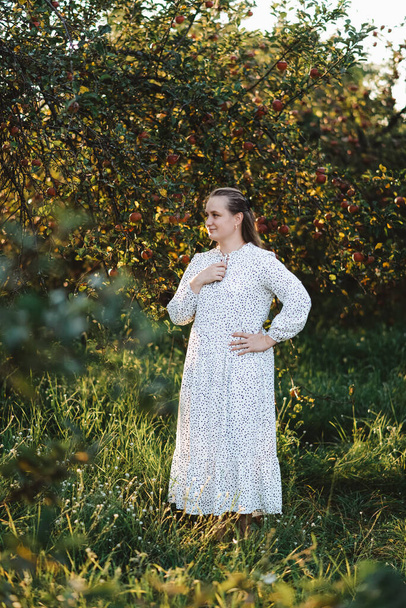 Elma bahçesinde uzun beyaz elbiseli genç bir kadın yan tarafa bakıyor. - Fotoğraf, Görsel