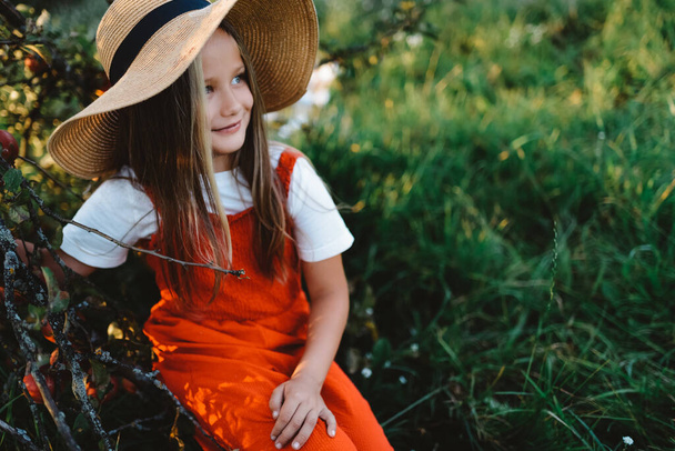 Dívka v klobouku a montérkách sedí na zahradě, usmívá se a dívá se na prázdný prostor - Fotografie, Obrázek