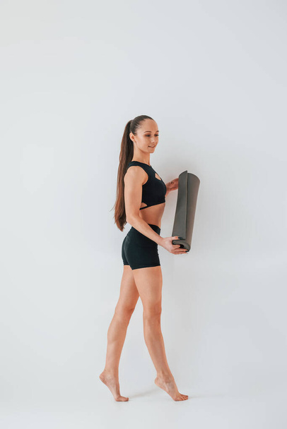 Standing and holding mat. Young woman in sportive clothes doing gymnastics indoors. - 写真・画像
