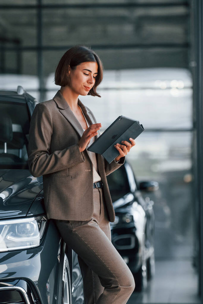 With tablet in hands. Woman is indoors near brand new automobile indoors. - Foto, immagini