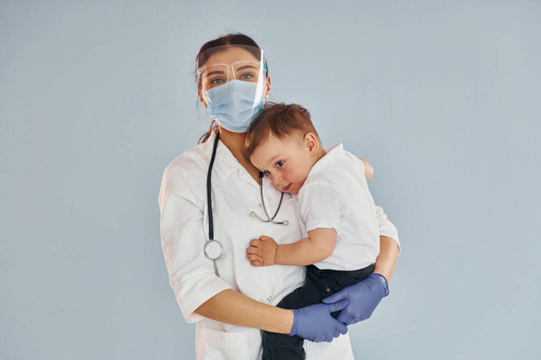 Νεαρή νοσοκόμα με λευκό παλτό και στηθοσκόπιο κρατά το αγοράκι στα χέρια. - Φωτογραφία, εικόνα