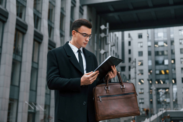 With notepad in hands. Businessman in black suit and tie is outdoors in the city. - Foto, afbeelding