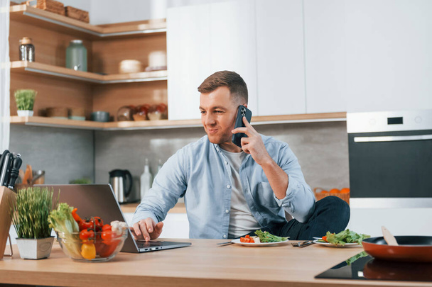 Talking by phone. Man preparing food at home on the modern kitchen. - Foto, afbeelding