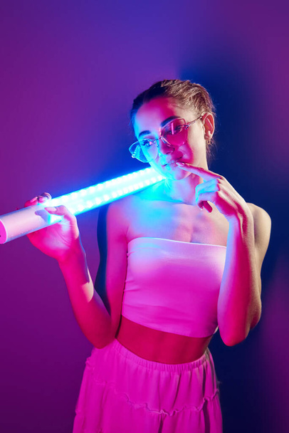 In glasses. Fashionable young woman standing in the studio with neon light. - Фото, изображение
