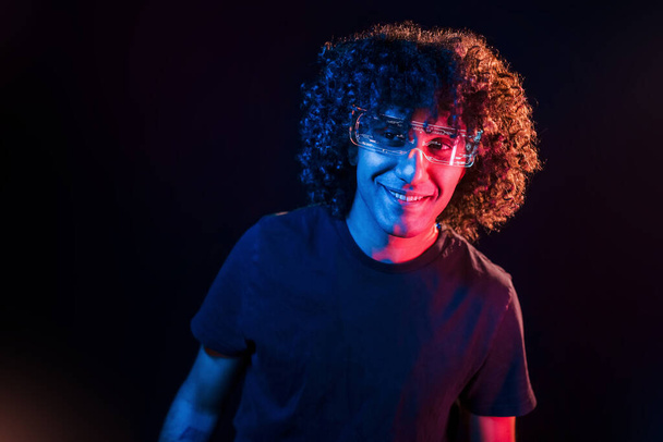 Young beautiful man with curly hair is indoors in the studio with neon lighting. - Photo, Image