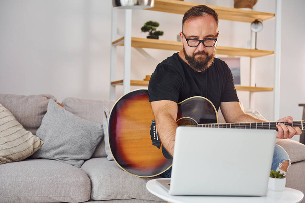 Using laptop. Man in casual clothes and with acoustic guitar is indoors. - 写真・画像