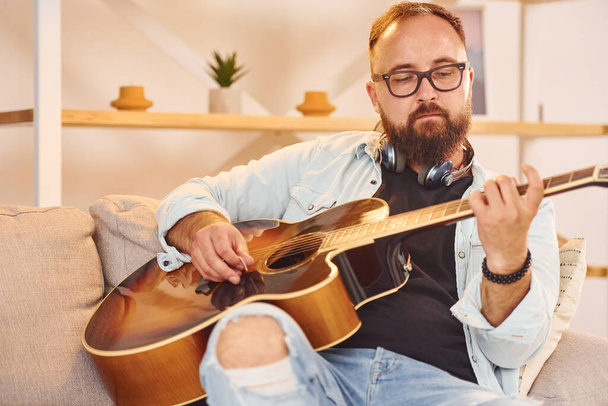 Creative occupation. Man in casual clothes and with acoustic guitar is indoors. - Foto, Bild