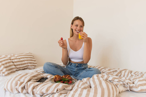 Smiling woman holding cherry tomato and orange juice near croissants on bed  - Foto, Imagem