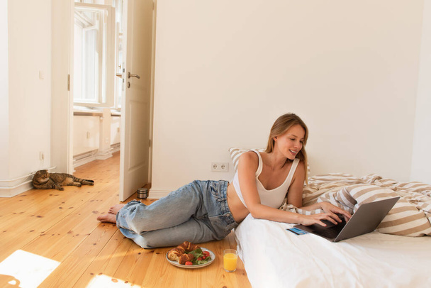 Mujer sonriente utilizando el ordenador portátil cerca de la tarjeta de crédito y desayuno en el dormitorio  - Foto, Imagen