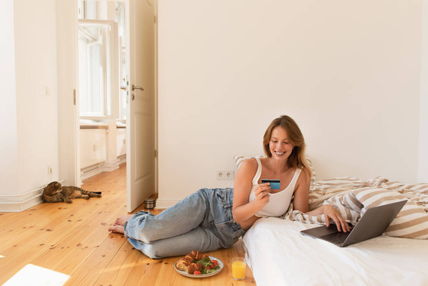 Mujer joven usando portátil y tarjeta de crédito cerca del desayuno y gato pliegue escocés en casa  - Foto, imagen
