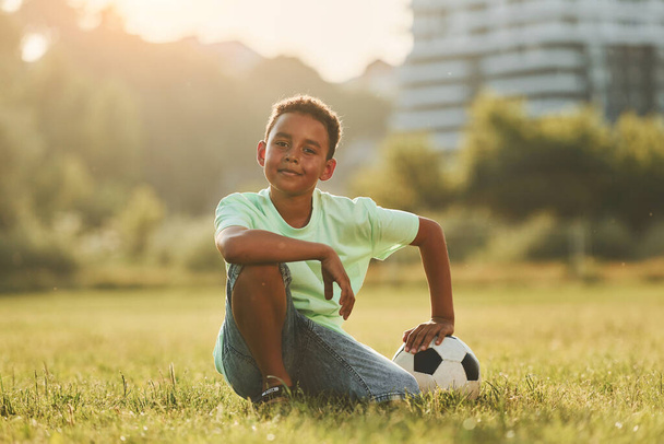 Taking a break. With soccer ball. African american kid have fun in the field at summer daytime. - Foto, afbeelding