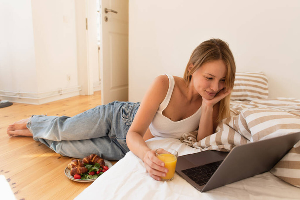 Smiling freelancer looking at laptop and holding orange juice near tasty breakfast on floor at home  - 写真・画像