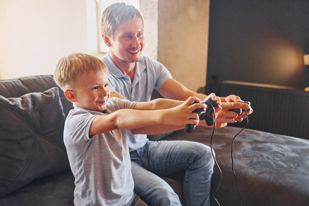 With video game joysticks. Father and son is indoors at home together. - Fotó, kép