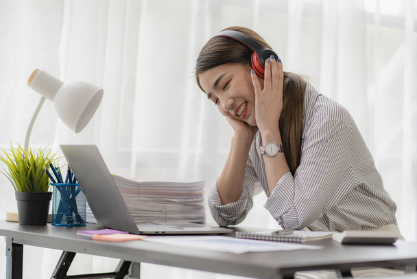 Egy ázsiai könyvelő vagy irodai dolgozó fejhallgatót hord online laptoppal, és szünetek alatt zenét hallgat.. - Fotó, kép